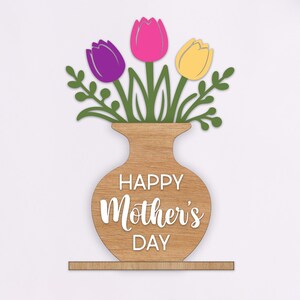 Può includere: Decorazione in legno per la festa della mamma. Un vaso marrone chiaro con tre tulipani viola, rosa e gialli. Il vaso ha le parole "HAPPY Mother's DAY" in bianco. Decorazione da esporre.