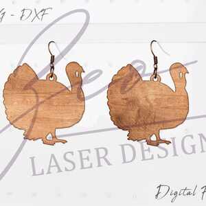 May include: A pair of wooden turkey earrings with a brown wood grain finish. The earrings are shaped like a turkey with a rounded body and a long neck. The earrings are hanging from a silver hook.