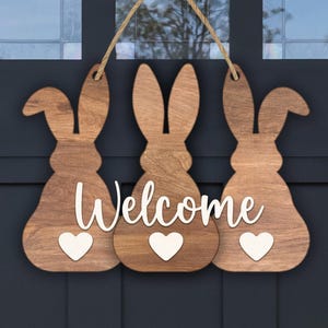 May include: Wooden welcome sign featuring three bunny silhouettes with white hearts. The word "Welcome" is in white script across the center. The sign is made of wood and hangs from a natural twine rope, perfect for Easter or spring decor.
