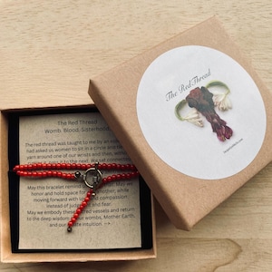 May include: A red beaded bracelet with a toggle clasp, displayed in an open brown box with a black interior. The box also contains a card with text about the red thread. A round sticker on the box reads "The Red Thread."