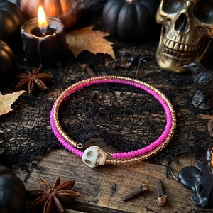 Könnte beinhalten: Ein zweireihiges Wickelarmband bestehend aus einer Reihe leuchtend pinkfarbener Saatperlen und einer Reihe metallisch goldener Saatperlen, die durch eine kleine, cremeweiße, geschnitzte Totenkopfperle in der Mitte verbunden sind.
