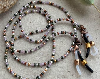 Puede incluir: Una cadena de gafas con cuentas con una mezcla de colores que incluyen morado, dorado, negro y plateado. La cadena tiene un diseño delicado y está hecha de pequeñas cuentas. La cadena tiene bucles de goma negra para sujetar las gafas.