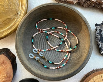 May include: A beaded eyeglass chain with white, black, red, and turquoise beads. The chain is resting in a dark wooden bowl. The chain has gold-colored metal clasps and is designed to hold eyeglasses.