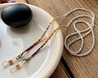 Op de afbeelding: Een glazen ketting met een mix van amberkleurige, bruine en zilveren kralen. De ketting heeft goudkleurige sluitingen en is ontworpen om een bril te houden. De ketting wordt gepresenteerd op een wit bord, met een zwarte steen.