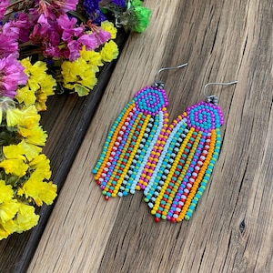 Puede incluir: Pendientes de cuentas coloridas con diseño de flecos. Los pendientes presentan una vibrante mezcla de colores, incluyendo turquesa, amarillo, naranja, rosa y blanco. Los pendientes se exhiben sobre una superficie de madera, con flores secas en el fondo.