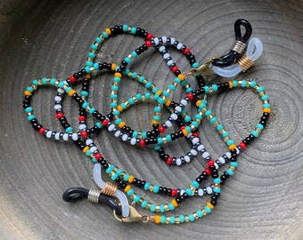 May include: A beaded eyeglass chain with black, white, red, gold, turquoise, and orange beads. The chain has black rubber loops to attach to glasses and gold-colored metal clasps. The chain is displayed in a wooden bowl.