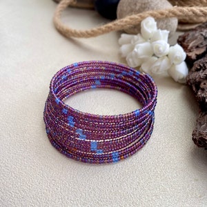 May include: A beaded bracelet with a spiral design. The bracelet is primarily purple with accents of blue beads. The bracelet is made of many strands of beads wrapped together. The bracelet is on a neutral background.