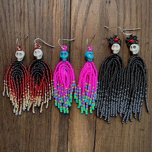 May include: Three pairs of beaded fringe earrings featuring skull beads. The first pair has red, black, and gold beads. The second pair has bright pink and turquoise beads. The third pair has black beads with red accents and white skull beads.