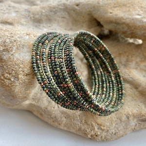 May include: A set of beaded bracelets in green, black, and copper hues. The bracelets are constructed from small, round beads and are arranged in a spiral. The bracelets are displayed on a light-coloured, textured surface.