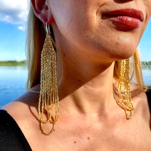 Puede incluir: Pendientes de cuentas doradas con un diseño en cascada. Los pendientes son largos y cuelgan del lóbulo de la oreja.