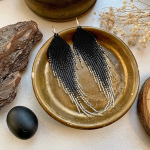 Puede incluir: Un par de pendientes largos de cuentas negras y plateadas. Los pendientes cuelgan de un plato dorado.