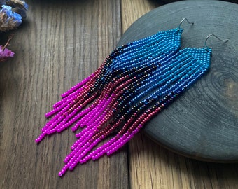 Könnte beinhalten: Ein Paar lange, geperlte Ohrringe mit einem Farbverlauf aus rosa, lila, blauen und schwarzen Perlen. Die Ohrringe hängen an silbernen Haken.