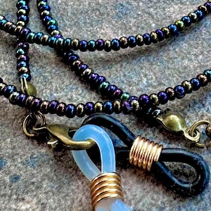 May include: A beaded glasses chain in blue, green, and purple hues. The chain is made of small, iridescent beads and has gold-coloured metal clasps and clear rubber loops to hold the glasses. The chain is designed to be worn around the neck.