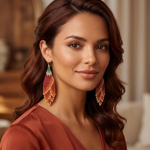 May include: A pair of long, diamond-shaped beaded earrings featuring a gradient pattern of deep red, orange, and yellow hues, finished with a small green section at the top. The earrings hang from gold-toned hooks and have a fringe-like bottom edge.