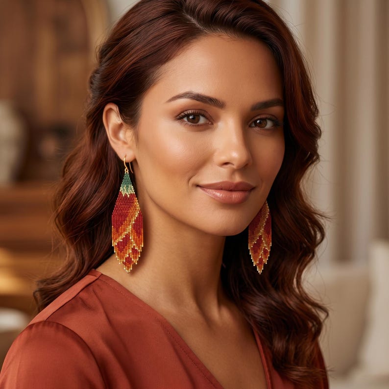 May include: A pair of long, diamond-shaped beaded earrings featuring a gradient pattern of deep red, orange, and yellow hues, finished with a small green section at the top. The earrings hang from gold-toned hooks and have a fringe-like bottom edge.