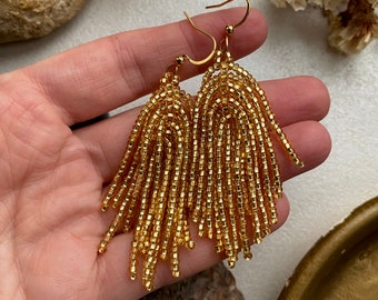 May include: A pair of gold-coloured beaded fringe earrings featuring a tiered arch design. Each earring is constructed from numerous small, metallic-finish seed beads arranged in vertical strands that hang from a simple gold-toned metal hook wire.