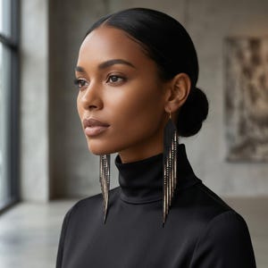 May include: Long, dangling earrings with a black, gold, and silver beaded design. The earrings are worn by a person wearing a black turtleneck. The earrings have a fringe-like appearance, with multiple strands of beads hanging down.
