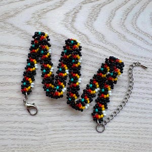 May include: A beaded bracelet with a black base, accented with red, yellow, white, and turquoise beads. The bracelet has a silver-tone clasp and an adjustable chain. The beads are tightly woven together.