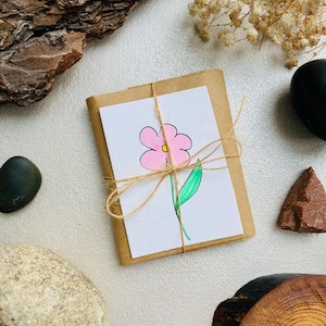 May include: A small, wrapped gift with a hand-drawn pink flower on a white card, tied with twine. The gift is on a white surface, surrounded by decorative elements like rocks, wood bark, and dried flowers.