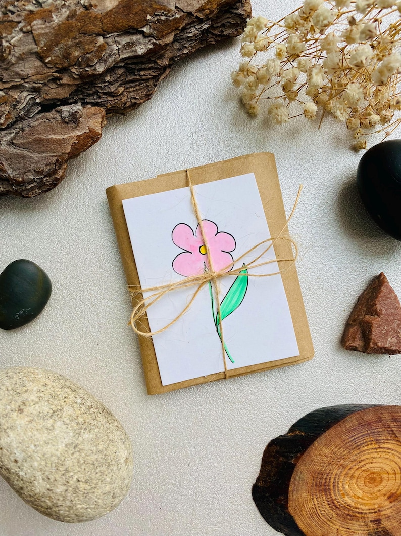 May include: A small, wrapped gift with a hand-drawn pink flower on a white card, tied with twine. The gift is on a white surface, surrounded by decorative elements like rocks, wood bark, and dried flowers.