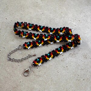 May include: A beaded bracelet with a black base, featuring a repeating pattern of red, yellow, white, and turquoise beads. The bracelet has a silver-colored chain and clasp for adjustable sizing. The beads are small and tightly woven.