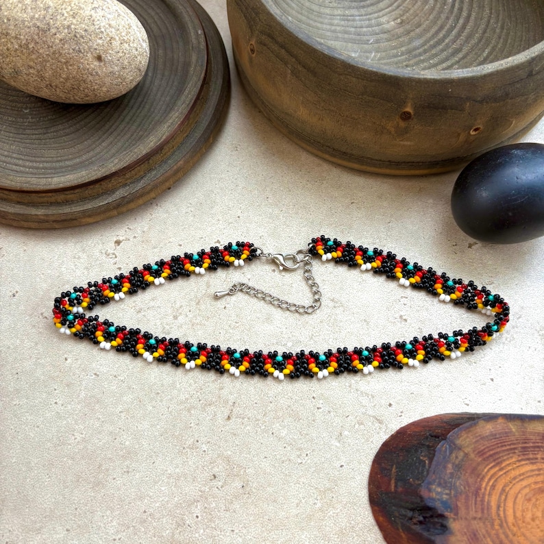 May include: A beaded choker necklace with a black base and colorful accents of red, yellow, white, and turquoise. The necklace has a silver-tone clasp and is displayed on a neutral surface. The necklace is approximately 14 inches long.