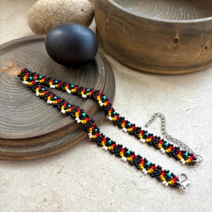 May include: Two beaded bracelets with a repeating pattern of black, red, yellow, white, and turquoise beads. The bracelets have a silver-colored chain and clasp. The bracelets are displayed on a wooden surface.