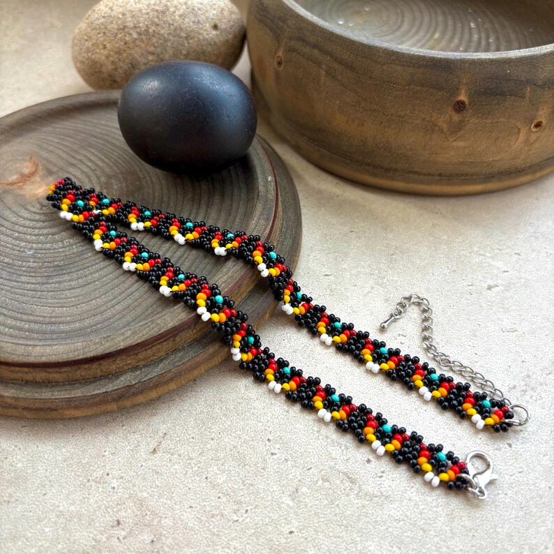May include: Two beaded bracelets with a repeating pattern of black, red, yellow, white, and turquoise beads. The bracelets have a silver-colored chain and clasp. The bracelets are displayed on a wooden surface.