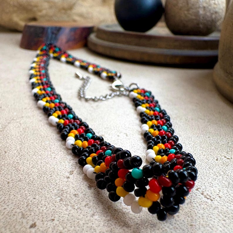 May include: A beaded necklace with a colorful pattern. The necklace features black, white, red, yellow, and turquoise beads. The necklace has a silver-colored chain clasp.
