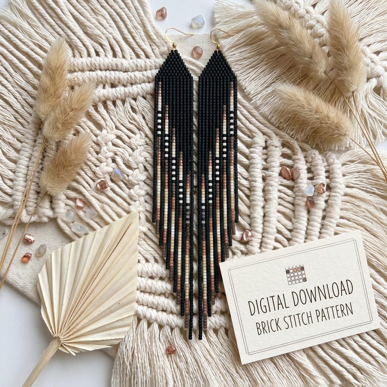 May include: Black beaded earrings with a geometric pattern in white, tan, and brown. The earrings are long and dangle, with gold-colored hooks. The background includes dried plants and a card that says "DIGITAL DOWNLOAD BRICK STITCH PATTERN."