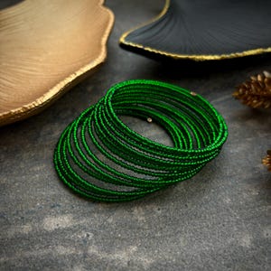 Puede incluir: Un conjunto de pulseras de cuentas verdes. Las pulseras están hechas de pequeñas cuentas redondas que están enhebradas en un alambre. Las pulseras están apiladas una encima de la otra, creando un aspecto grueso y en capas.