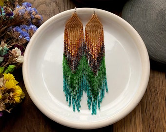 Op de afbeelding: Een paar lange, geperde oorbellen met een kleurverloop van goud naar groen naar turquoise. De oorbellen hangen op een wit bord.
