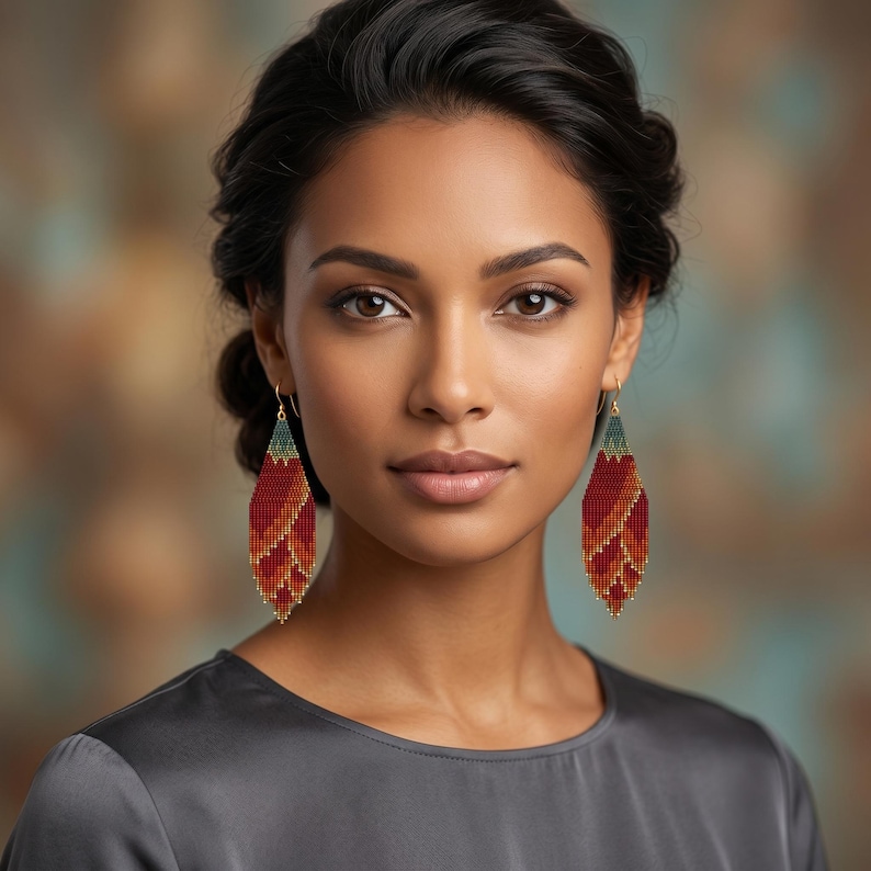 May include: A pair of long, diamond-shaped beaded earrings featuring a gradient pattern of deep red, orange, and yellow hues, transitioning to a dark teal at the top. The earrings are suspended from thin gold-toned metal hooks.