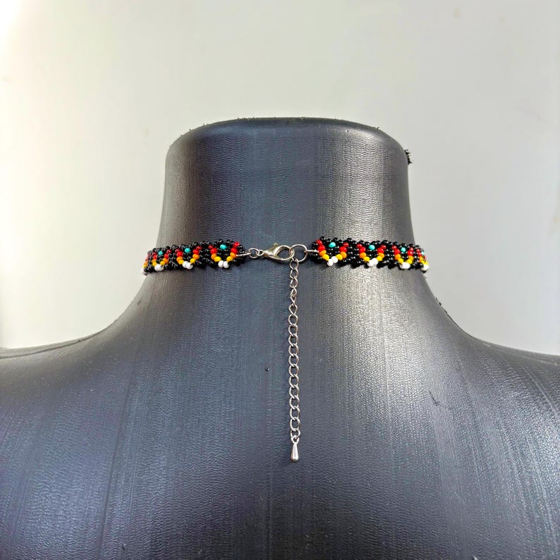 May include: Black beaded choker necklace with a colorful pattern of red, yellow, and turquoise beads. The necklace has a silver-colored clasp and an extender chain. The choker is displayed on a black mannequin.