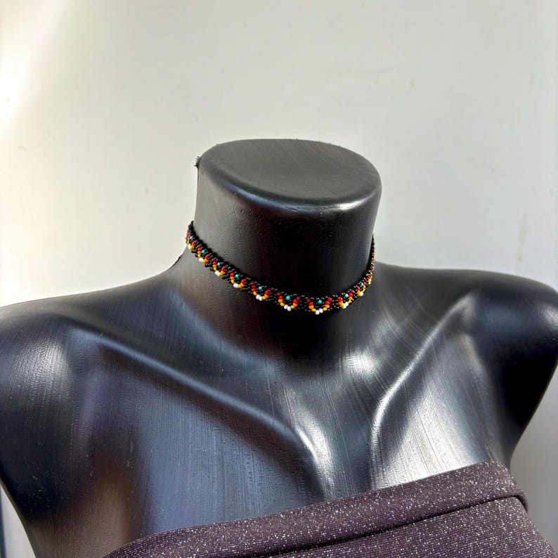 May include: A black beaded choker necklace with a scalloped design. The necklace features orange, white, and green beads. The necklace is displayed on a black mannequin.