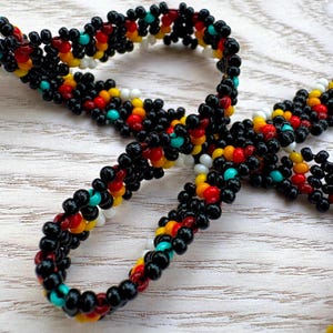 May include: A beaded necklace with a repeating pattern of black, red, yellow, white, and turquoise beads. The necklace is made of small, round beads strung together to form a flexible chain. The beads are arranged in a symmetrical design.