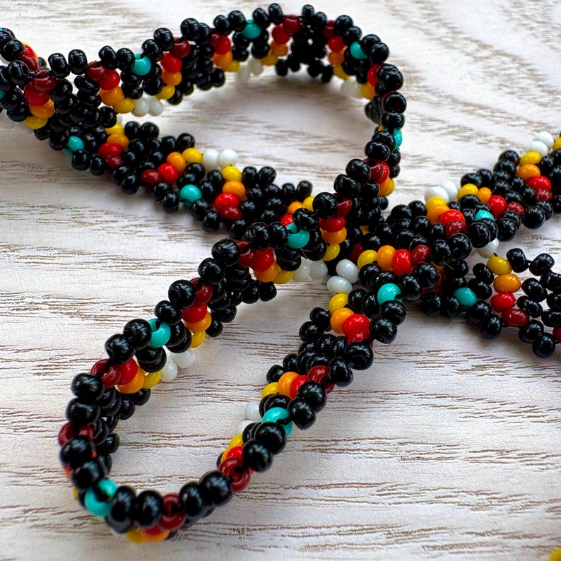 May include: A beaded necklace with a repeating pattern of black, red, yellow, white, and turquoise beads. The necklace is made of small, round beads strung together to form a flexible chain. The beads are arranged in a symmetrical design.