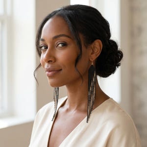May include: A woman wearing long, black beaded earrings with gold accents. The earrings have a rectangular top and multiple strands of beads that hang down. The woman is wearing a cream-colored top.