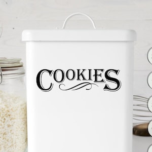 Op de afbeelding: Witte metalen koekjesbus met zwarte tekst die "COOKIES" zegt met een decoratieve versiering. De bus staat op een wit oppervlak met een pot witte vlokken, een kom havermout en een garde.