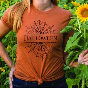 May include: A woman wearing an orange t-shirt with a black spiderweb design and the text "This is HALLOWEEN".