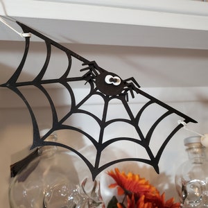 Puede incluir: Corte de papel negro de una telaraña con una araña negra con ojos saltones blancos colgando de una cuerda blanca. La telaraña está colgada de una estantería blanca.