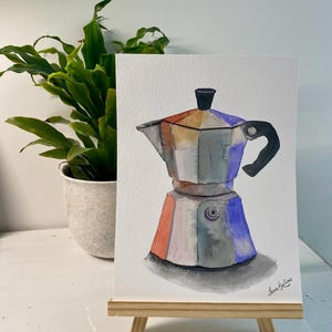 Pode incluir: Pintura em aquarela de uma cafeteira italiana prateada e colorida. A pintura apresenta uma alça e uma parte superior pretas, com tons de laranja, azul e cinza. A obra de arte é exibida em um pequeno cavalete de madeira.