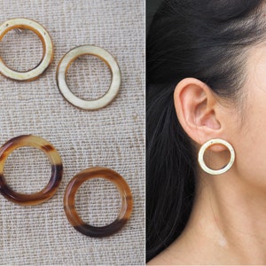May include: A pair of brown and white hoop earrings. The earrings are made from a natural material, possibly horn or bone. The earrings are shown on a model's ear.