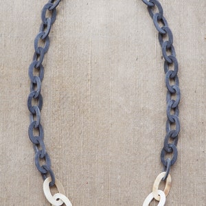May include: A necklace featuring a chain design. The upper portion of the chain is a dark gray color, transitioning to a cream color at the bottom. The oval links are connected to form a necklace.