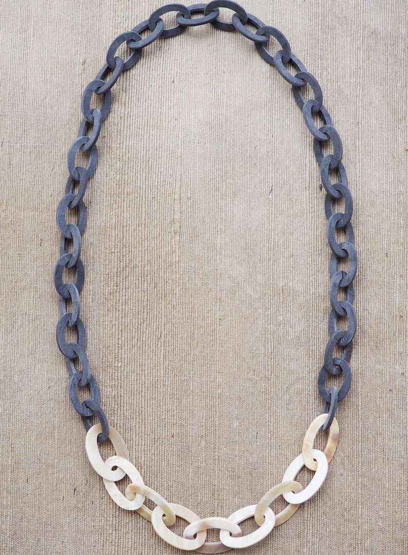 May include: A necklace featuring a chain design. The upper portion of the chain is a dark gray color, transitioning to a cream color at the bottom. The oval links are connected to form a necklace.