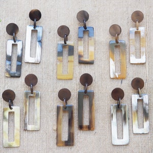 May include: A collection of rectangular drop earrings with a circular top. The earrings feature a hollowed-out rectangle design with a mix of light and dark brown, and white tones. The earrings are displayed on a neutral background.