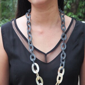 May include: A long chain necklace with large oval links. The upper portion of the chain is a dark gray color, transitioning to a cream color at the bottom. The necklace is worn over a black top.