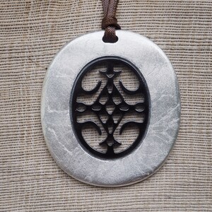 May include: A silver pendant with a black, intricate design in the center. The pendant is oval-shaped and hangs from a brown cord.