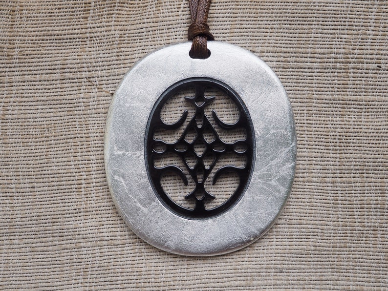 May include: A silver pendant with a black, intricate design in the center. The pendant is oval-shaped and hangs from a brown cord.