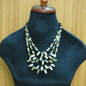 May include: A multi-strand necklace with oval-shaped beads in shades of cream, brown, and black. The necklace is displayed on a black mannequin bust.
